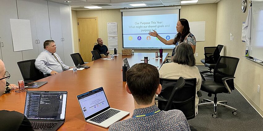 woman stands and talks to group of people seated at a conference table