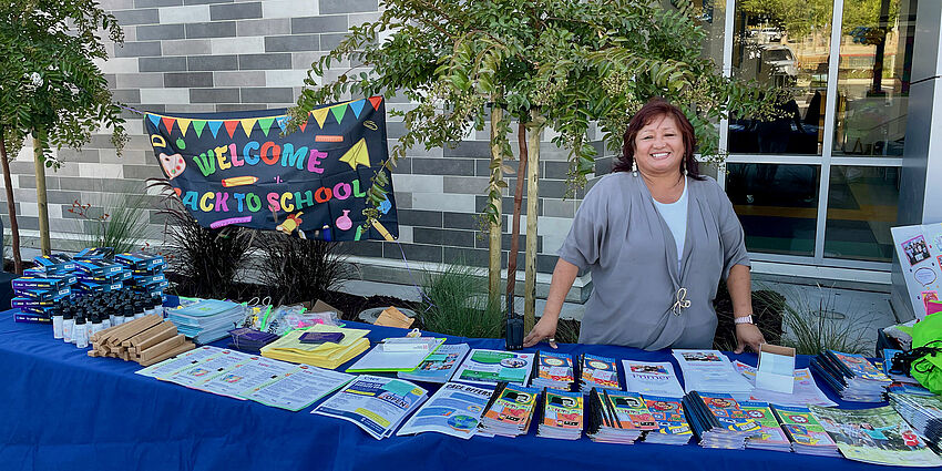 a woman smiles while standing at a table filled with helpful items for families. Sign behind her says Welcome back to School