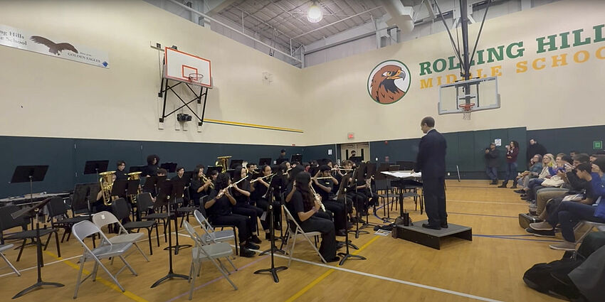 A school band performs in a gymnasium with a conductor leading. Students play instruments while an audience watches.