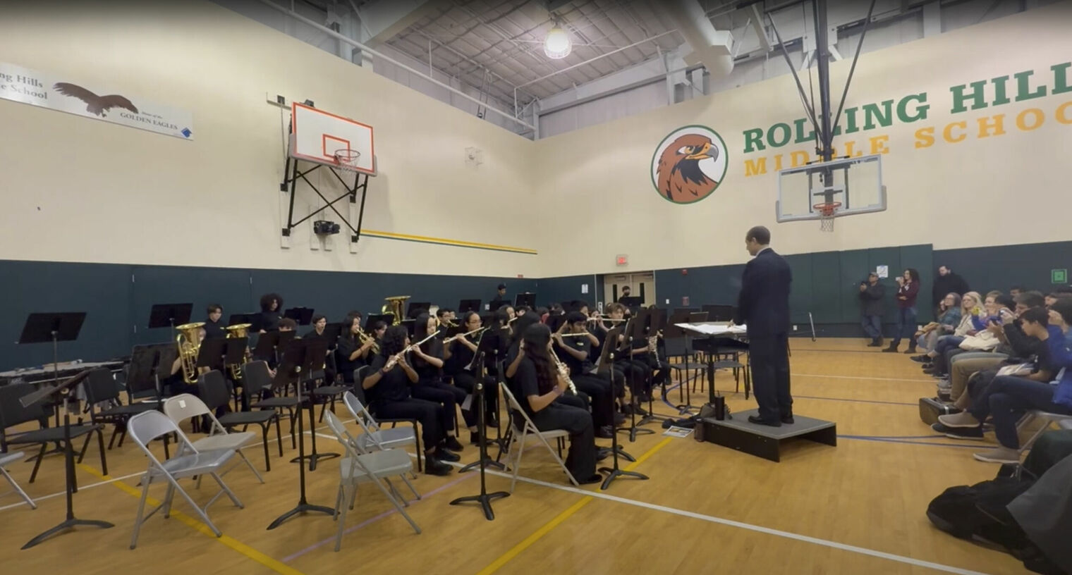 A school band performs in a gymnasium with a conductor leading. Students play instruments while an audience watches.